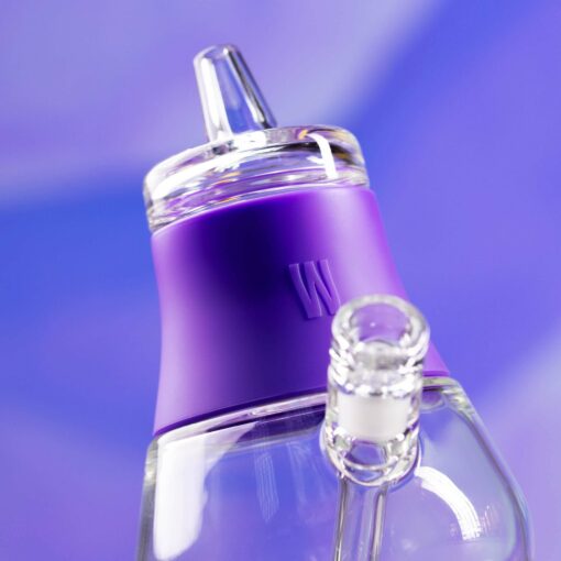A close-up of The Bubbler - Grape glass bottle with a purple silicone sleeve and spout, set against a blue and purple blurred background. The sleeve displays an embossed W logo.