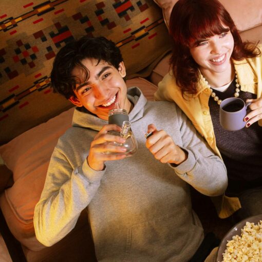 Two young adults relax on a couch, smiling. One holds The Bubbler - Smoke and a lighter, while the other enjoys a mug. A bowl of popcorn sits on the table as they enjoy their smoke session together.