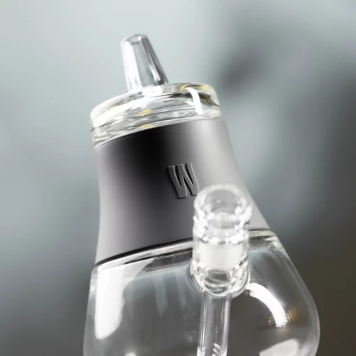 Close-up of The Bubbler - Smoke, a modern glass smoking accessory with a black neck band and a raised W logo, set against a blurred gray background.