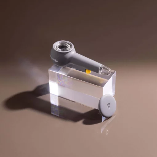 The Pipe - Smoke is a modern gray pipe with a transparent mouthpiece, resting on a clear block. A matching round lid is nearby, all displayed against a smooth, neutral background. Subtle smoke is suggested by its sleek design.