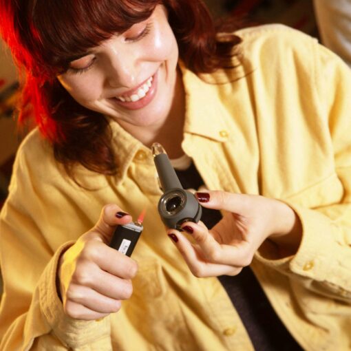 A smiling person with red hair in a yellow shirt holds a lighter in one hand and gets ready to use The Pipe - Smoke in the other.