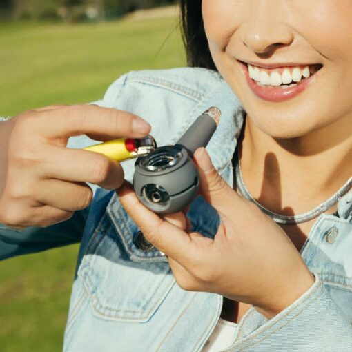 A smiling person outdoors in a light denim jacket and silver necklace holds The Pipe - Smoke, lighting it against a sunlit, grassy background.