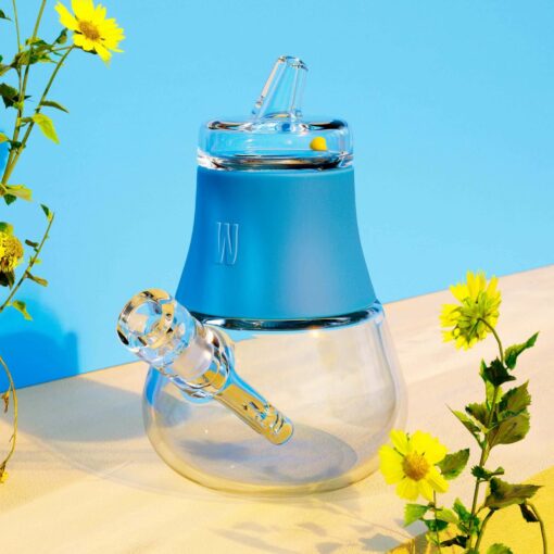 The Pipe - Hot Pink is a modern glass water pipe with a bold hot pink silicone top, shown on a light surface with yellow flowers and bright blue background accents.