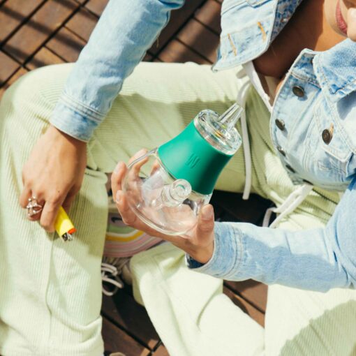 Seated cross-legged in a forest, a person in light green corduroy pants and a denim jacket holds The Bubbler - Forest in one hand and a yellow lighter in the other.