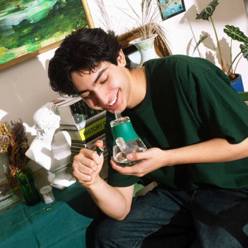A person in a green shirt uses The Bubbler - Forest, holding a lighter. Seated at a table with books, a white bust, and plants, they enjoy the vibrant Forest-themed room filled with unique decor.