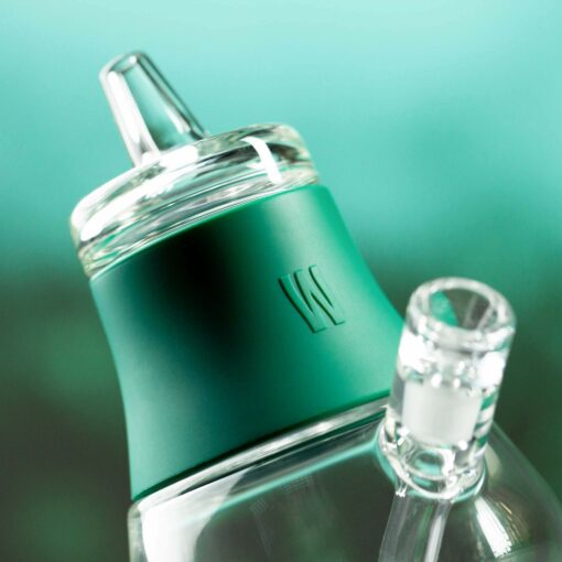 Close-up of The Bubbler - Forest, a glass bubbler bottle with a green silicone band featuring a raised W logo and a clear spout, set against a blurred teal forest background.