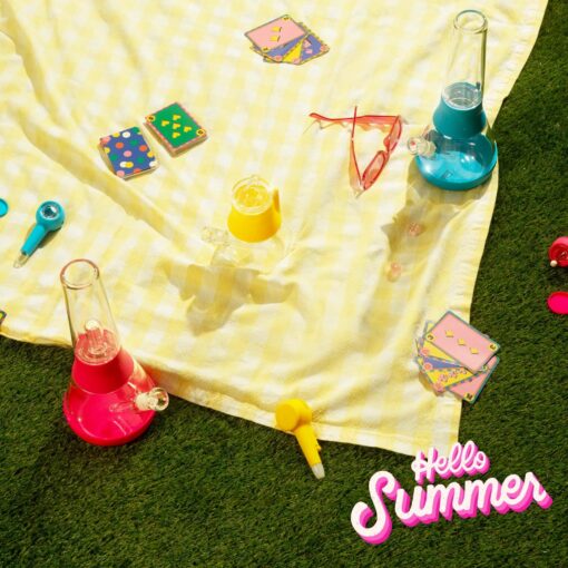 A yellow picnic blanket on grass with colorful glass bongs, The Pipe - Hot Pink, sunglasses, playing cards, and “Hello Summer” in hot pink and white script in the bottom right corner.