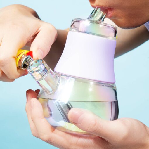 Someone uses The Iridescent Bubbler - Lavender, holding its small, transparent iridescent body with a lavender accent, lighting it with a yellow lighter and placing their mouth on the mouthpiece against a light blue background.