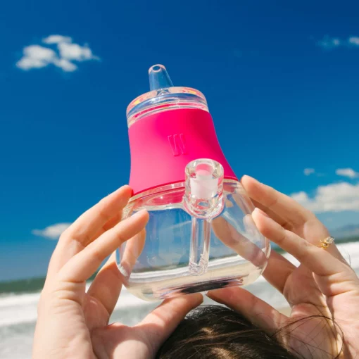 At the beach under a bright sky, someone holds The Bubbler - Hot Pink, a clear glass bubbler with a vibrant silicone top—an eye-catching smoking accessory perfect for sunny days by the ocean.