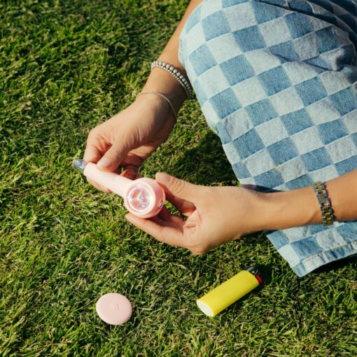 A person in blue checkered pants and silver bracelets sits on grass, holding The Pipe Iridescent - Bubblegum with a clear cap. Nearby are a yellow lighter and a round pink lid on the ground.