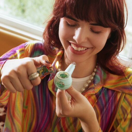 A smiling person with red hair and a colorful striped shirt lights The Pipe Iridescent - Sky indoors, holding it in one hand and a lighter in the other.