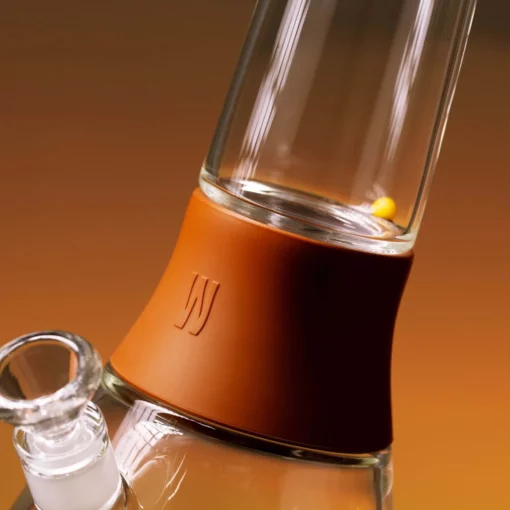 A close-up of The Bong - Leather, a glass water pipe with a brown silicone band around the neck and an embossed W logo. The background features a smooth gradient of warm brown and orange hues.