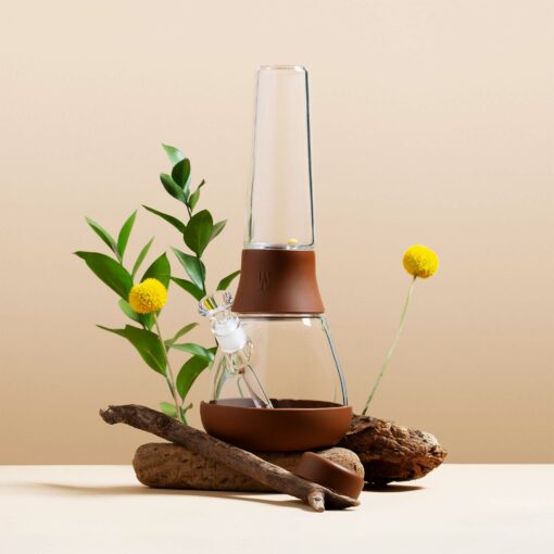 The Bong - Leather is showcased among green leaves, yellow billy ball flowers, and rustic wooden branches on a beige background—offering a stylish twist on the classic bong design.
