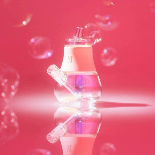 The Bubbler Iridescent - Bubblegum, a compact transparent glass bubbler with a pink silicone base, rests on a reflective pink surface surrounded by floating bubbles and glowing softly in pink light.