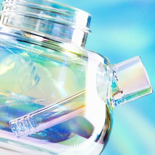 Close-up of The Bubbler Iridescent - Bubblegum: a clear glass jar with spout and straw, shining with iridescent colors against a soft blue-green blur for a dreamy, abstract look.