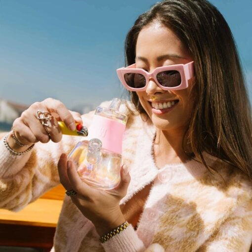 A smiling woman in pink sunglasses and a patterned sweater lights The Bubbler Iridescent - Bubblegum outdoors on a sunny day.