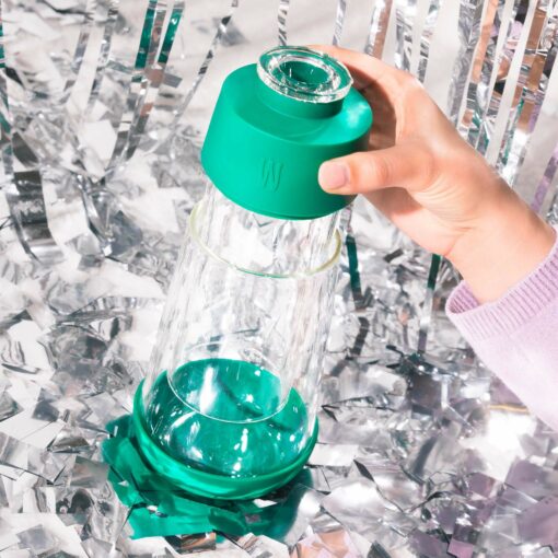 A hand holds The Gravity Bong - Forest, a clear glass bottle with a green silicone lid and base, surrounded by shiny silver streamers that evoke the fresh hues of a forest.