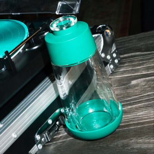 The Gravity Bong - Forest, a clear glass tumbler with teal silicone top and bottom, sits at an angle on wood beside a teal turntable and silver case, creating a modern, forest-inspired vibe.