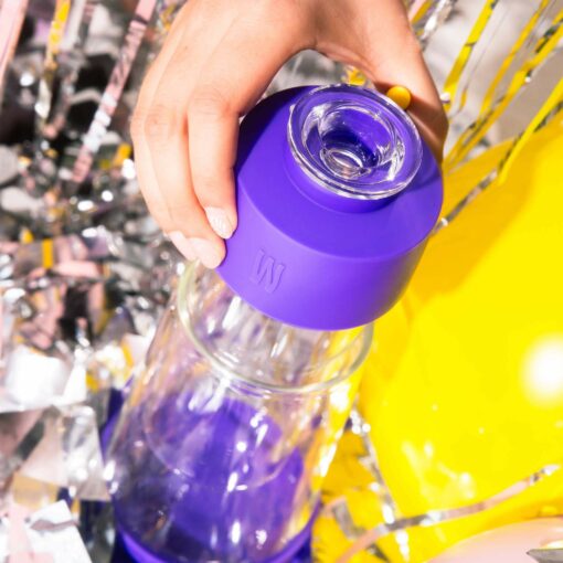 A hand holds the cap of The Gravity Bong - Grape, a clear glass water bottle with a purple, grape-inspired lid, surrounded by shiny silver and yellow decorations.