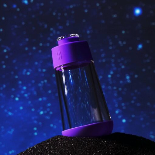 The Gravity Bong - Grape features a clear bottle with grape-purple lids, balanced on a textured black surface against a starry night sky, capturing the playful spirit of a gravity bong.