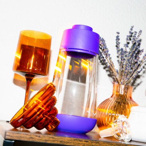 The Gravity Bong - Grape, featuring a purple cap and grape hue, stands on a wooden surface next to an amber wine glass, an amber vase with lavender stems, and a honey dipper under bright lighting.