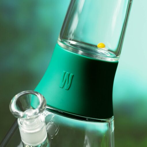 Close-up of The Bong - Forest: a glass bong with a green silicone band embossed with a W logo and a yellow bead inside the chamber. The blurred teal and green background evokes peaceful forest hues.