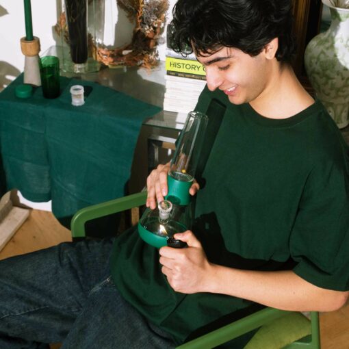 A young person with dark hair sits indoors in a green chair, smiling and holding The Bong - Forest. Nearby, forest-green and neutral objects, a book titled HISTORY, and potted plants are on a green-draped table.