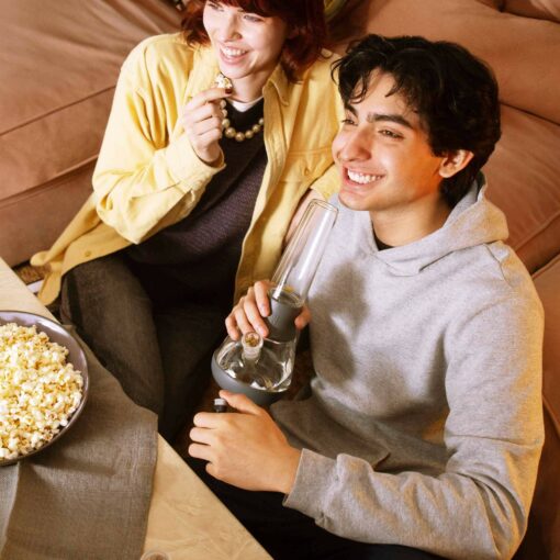 Two people relax on a couch with popcorn. One enjoys a snack while the other smokes from The Bong - Smoke, sharing a laid-back moment together.