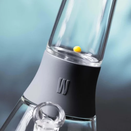 Close-up of The Bong - Smoke, a glass water pipe with a gray silicone neck band and raised W logo. A small yellow ball sits inside. The blurred blue background highlights the smoke experience.
