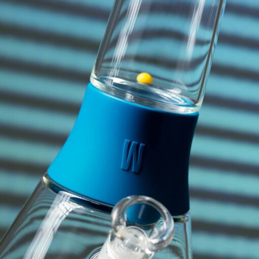 Close-up of The Bong - Midnight, a glass bong with a blue silicone neck band and a yellow marble inside. The blurred background shows midnight blue and black horizontal stripes.