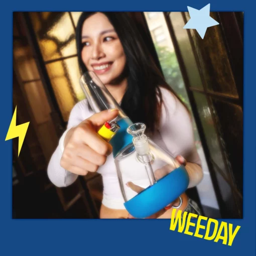 A smiling woman holds The Bong - Midnight, a blue and clear glass piece, with a lighter. The image has a blue border featuring a star, lightning bolt, and bold yellow “WEEDAY,” giving off midnight vibes.