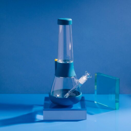 The Bong - Midnight, a modern glass bong with blue accents, sits on a blue platform against a midnight blue background beside a transparent blue-green glass block in a brightly lit, minimalistic scene.
