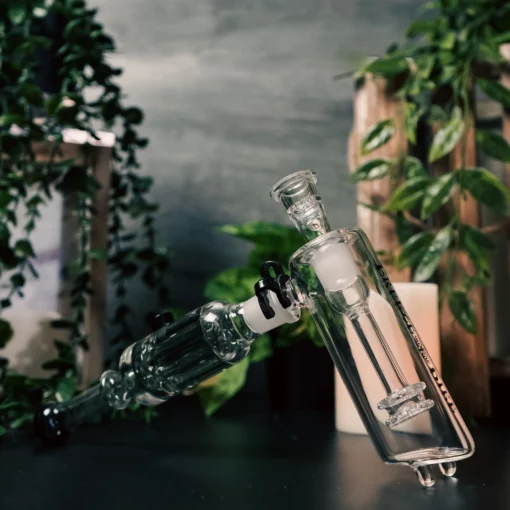A close-up of the Freeze Pipe Bubbler Pro with detailed parts rests on a dark surface, while green leafy plants, wood accents, and a white candle in the background add to the calm indoor vibe.