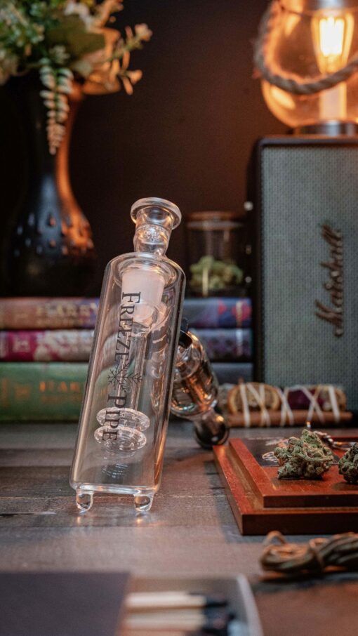 A Freeze Pipe Bubbler Pro sits on a table with cannabis buds, books, and a lit lamp in the background, creating a cozy and relaxed vibe.