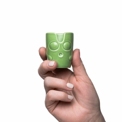 A hand with light pink nails holds a small green ceramic cup with a stylized face—part of the BigFUN! Potheads Ashtray Set, perfect for quirky decor lovers. The scene is set against a white background.
