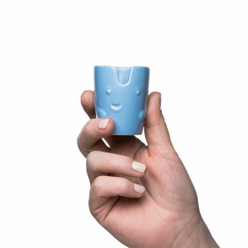 A hand with manicured nails holds a light blue BigFUN! Potheads Ashtray Set cup featuring a smiling face with raised eyes and cheeks, set against a plain white background.