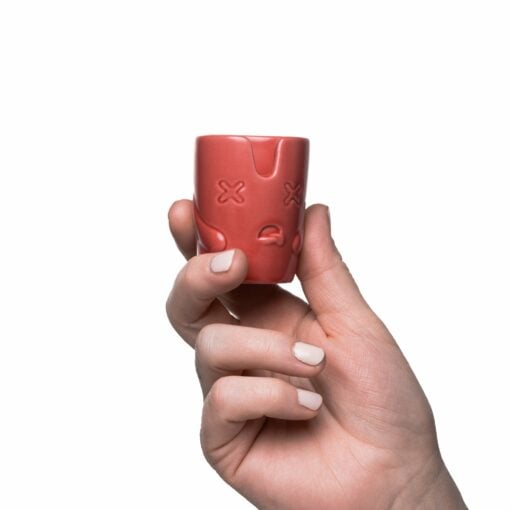 A hand with light-colored nail polish holds a small pink ceramic cup from the BigFUN! Potheads Ashtray Set, featuring an embossed cartoon face with X-shaped eyes and a slight frown, set against a plain white background.
