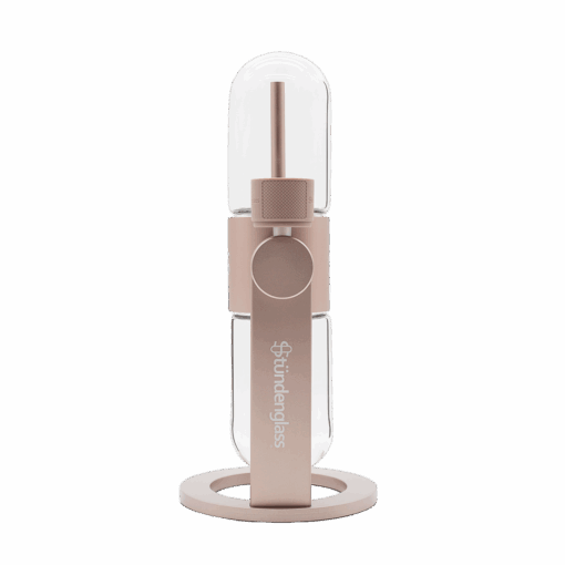 The Stündenglass Gravity Infuser - Desert Rose | 360° Gravity Hookah features a modern cylindrical glass and metallic design with a rose gold finish, clear chamber, and logo, standing upright on a circular base against a white background.